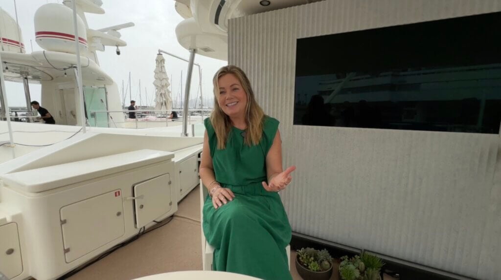 A woman in a green dress sits and gestures with one hand on the deck of a yacht, her pose capturing a sense of human touch, with marina boats visible in the background.